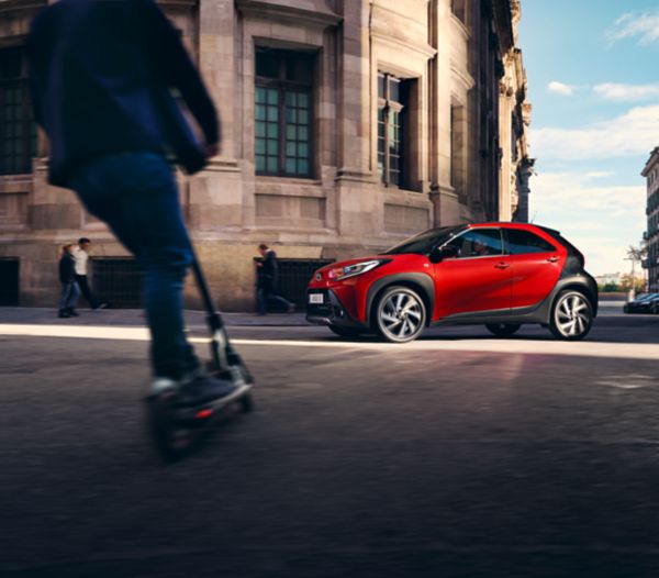 Image showing Chili Red Aygo X driving in a city street with scooter rider approaching in the foreground