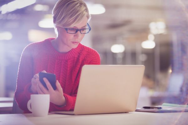 A lady wearing glasses and a red jumper looks at her laptop with her smartphone in her hand. A coffee cup sits on the table.