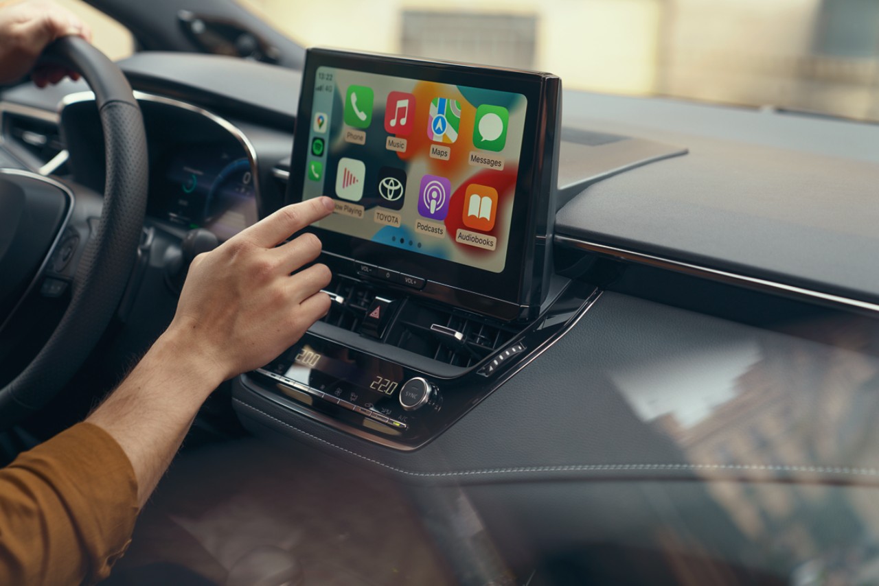 A person inside a Toyota interacts with the car's multimedia touch screen. The display shows the Apple CarPlay home screen.