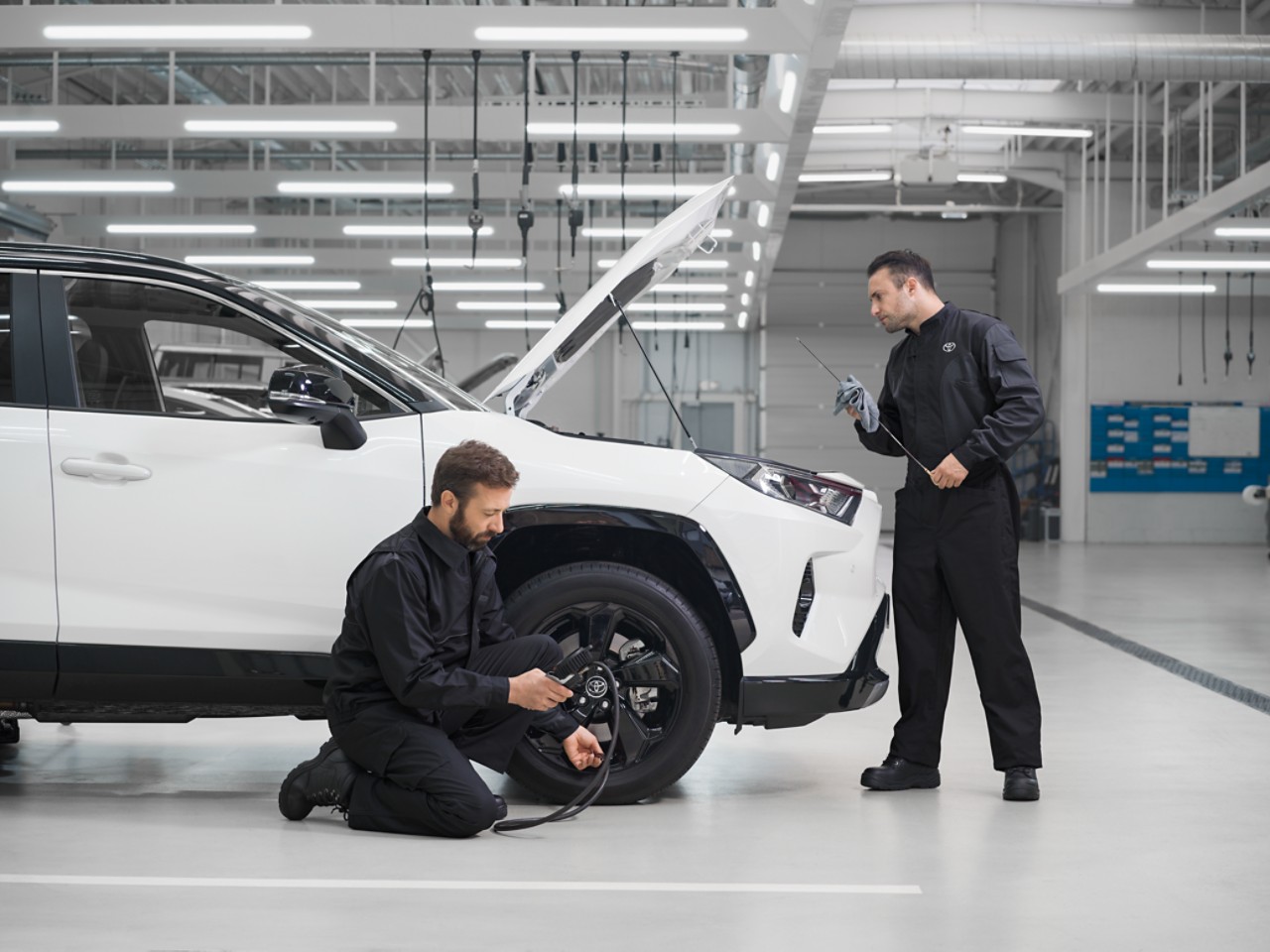 Two technicians service a white Toyota RAV4, its bonnet is up. One kneels down inspecting tyre pressure, the other oil level.